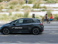 Ein schwarzes Sharetoo-Auto steht auf der Straße vor zwei sitzenden Personen